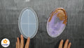 Zwei Hände halten ovale FLEURY Silikonform für Resin Art (AM1120) Formen auf einer grauen Fläche; die linke Form ist leer, die rechte zeigt lila und goldenes Harz. Das FLEURY Logo erscheint in der unteren linken Ecke.